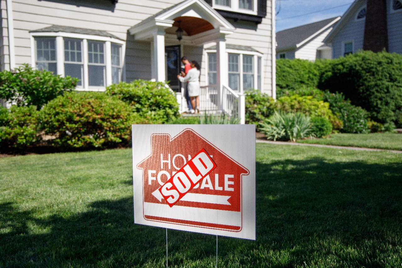 Sold home for sale sign in front yard with couple on front porch